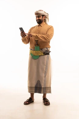 Using modern technologies in daily life: making video calls, chatting with friends, browsing websites and internet applications. Portrait of a Yemeni Arab man wearing a shirt and izar with a turban and jambiya, holding a mobile phone in his hand. Full-length portrait, white background.