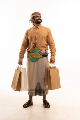 النظر الى شي ما، شراء الأغراض الجديدة و المستلزمات، الاستمتاع بالتسوق و الحصول على العروض , بورتريه لرجل عربي يمني يرتدي القميص والازار مع العمامة والجنبية يحمل بيديه أكياس ورقية , الجسم بالطول الكامل , خلفية بيضاء