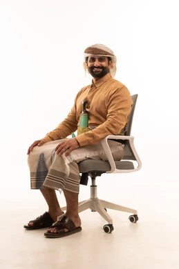 تمثيل الرجولة العربية الأصيلة، المحافظة على اللباس التقليدي،النظر الى الكاميرا تعابير السعادة و السرور، بورتريه لرجل عربي يمني يرتدي القميص والازار مع العمامة والجنبية يجلس على كرسي، خلفية بيضاء