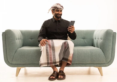 دمج التقنيات الحديثة فى الحياة اليومية، استخدام الهاتف المحمول ، تصفح مواقع التواصل الاجتماعي ، بورتريه لرجل عربي يمني يرتدي القميص والازار مع العمامة يجلس على الاريكة يمسك الجوال بيده ، خلفية بيضاء