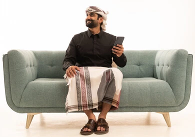 النظر الى شيء ما بتعابير السعادة، استخدام الهاتف المحمول ، دمج التقنيات الحديثة فى الحياة اليومية، تصفح مواقع التواصل الاجتماعي ، بورتريه لرجل عربي يمني يرتدي القميص والازار مع العمامة يجلس على الاريكة يمسك الجوال بيده ، خلفية بيضاء