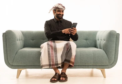 استخدام الهاتف المحمول ، دمج التقنيات الحديثة فى الحياة اليومية ، إجراء مكالمة هاتفية مع الأهل والأصدقاء، تصفح مواقع التواصل الاجتماعي ، بورتريه لرجل عربي يمني يرتدي القميص والازار مع العمامة يجلس على الاريكة يمسك الجوال بيده ، خلفية بيضاء