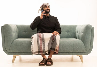 تبادل الأخبار مع الأهل والأصدقاء، إجراء مكالمة هاتفية باستخدام الموبايل، ورتريه لرجل عربي يمني يرتدي القميص والازار مع العمامة يجلس على الاريكة يمسك بيدية الجوال يقوم بالتواصل مع الأقارب، تعابير الفرح والسرور، خلفية بيضاء