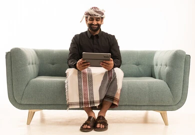 تعابير السعادة و السرور، استخدام التقنية الحديثه المتطورة في الحياة العملية عبر الجهاز اللوحي،  بورتريه لرجل عربي يمني يرتدي القميص والازار مع العمامة يجلس على الاريكة يقوم بالعمل على جهاز التابلت، خلفية بيضاء