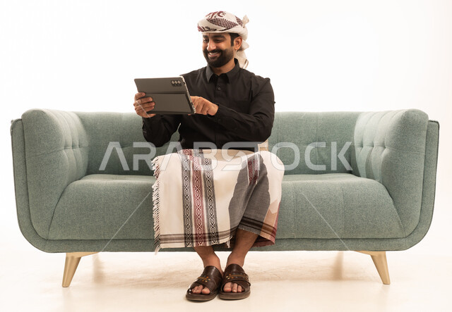 استخدام التقنية الحديثه المتطورة في الحياة العملية عبر الجهاز اللوحي، تعابير السعادة و السرور،  بورتريه لرجل عربي يمني يرتدي القميص والازار مع العمامة يجلس على الاريكة يقوم بالعمل على جهازالتابلت، خلفية بيضاء