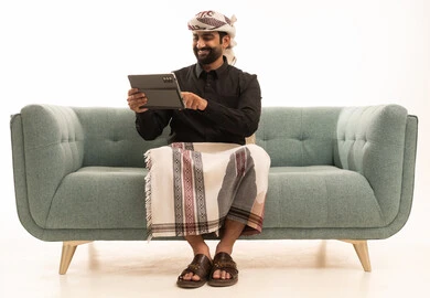 استخدام التقنية الحديثه المتطورة في الحياة العملية عبر الجهاز اللوحي، تعابير السعادة و السرور،  بورتريه لرجل عربي يمني يرتدي القميص والازار مع العمامة يجلس على الاريكة يقوم بالعمل على جهازالتابلت، خلفية بيضاء