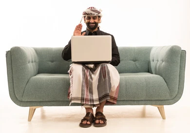 Making a video call, raising a hand with expressions of happiness and joy, completing work and tasks remotely via a laptop, a portrait of a Yemeni Arab man wearing a shirt and a waist wrap with a turban sitting on a sofa using a computer, white background.