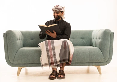 Spending time reading, learning, and culture, the concept of self-learning, expressions of contemplation and focus, a portrait of an Arab Yemeni man wearing a shirt and a sarong with a turban sitting on a sofa reading a book, white background.