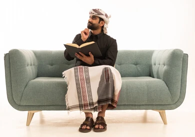 Spending free time on learning and culture, the concept of education and scientific research, the hobby of reading and exploration, a portrait of an Arab Yemeni man wearing a shirt and a sarong with a turban sitting on a sofa holding a book and resting his hand on his chin looking at something with a thoughtful expression, white background.