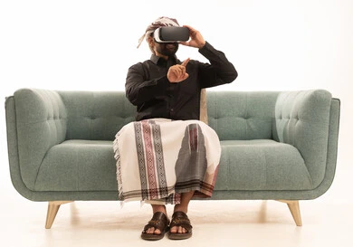 استخدام حاسة اللمس لملامسة عناصر هولوجرامية بإيماءات النقر, بورتريه لرجل عربي يمني يرتدي القميص والازار مع العمامة يجلس على الاريكة يرتدي نظارة الواقع الافتراضي ثلاثية الأبعاد يشير إلى شيء ما بأصبعه السبابة, محاكاة العالم الخيالي, خلفية بيضاء