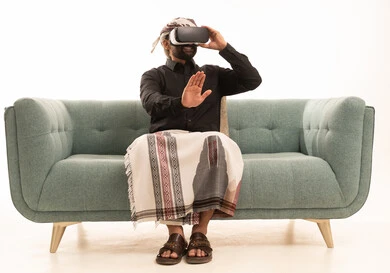 التطور التقني والتكنولوجي, المتعة و التسلية بالمشاهدة عبر نظارة الواقع الافتراضي، بورتريه لرجل عربي يمني يرتدي القميص والازار مع العمامة يجلس على الاريكة ينظر من خلال نظارات ثلاثية الأبعاد (الفي ار), الاندماج ومحاكاة العالم الخيالي والنظر الى الافلام الواقعيه، خلفيه بيضاء