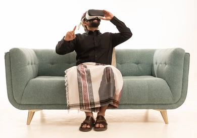 محاكاة العالم الخيالي، استخدام حاسة اللمس لملامسة عناصر هولوجرامية بإيماءات النقر, بورتريه لرجل عربي يمني يرتدي القميص والازار مع العمامة يجلس على الاريكة يرتدي نظارة الواقع الافتراضي ثلاثية الأبعاد يشير إلى شيء ما بأصبعه السبابة, خلفية بيضاء