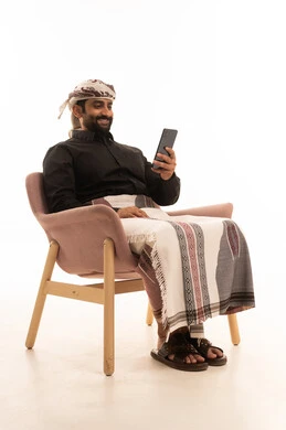The use of modern technological devices for entertainment and leisure, browsing social media, and a portrait of an Arab Yemeni man wearing a shirt and a sarong with a turban sitting on a chair holding a mobile phone in his hand, with a white background.