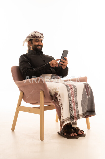 Browsing social media, using modern technological devices for entertainment and leisure, and a portrait of an Arab Yemeni man wearing a shirt and a sarong with a turban sitting on a chair holding a mobile phone in his hand, with a white background.