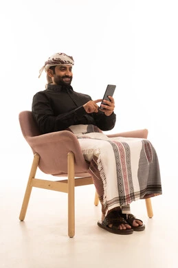Browsing social media, using modern technological devices for entertainment and leisure, and a portrait of an Arab Yemeni man wearing a shirt and a sarong with a turban sitting on a chair holding a mobile phone in his hand, with a white background. Browsing social media, using modern technological devices for entertainment and leisure, and a portrait of an Arab Yemeni man wearing a shirt and a sarong with a turban sitting on a chair holding a mobile phone in his hand, with a white background.