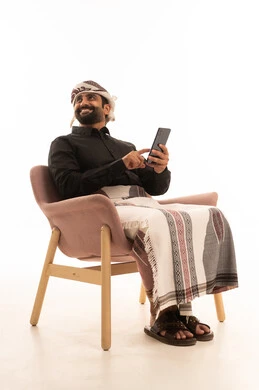 Looking at something, using modern technological devices for entertainment and amusement, browsing social media, and a portrait of an Arab Yemeni man wearing a shirt and a waist wrap with a turban sitting on a chair holding a mobile phone in his hand, with a white background.