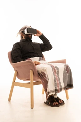 Using a modern and advanced technical device for entertainment and amusement, gestures of immersion and simulation of the imaginary world, wearing 3D VR glasses, a portrait of a Yemeni Arab man wearing a shirt and a waist wrap with a turban sitting on a chair looking through virtual reality glasses, white background.