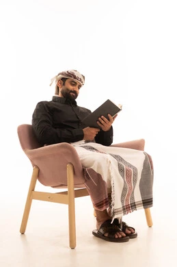 Expressions of contemplation and focus, the concept of self-learning, spending time reading, learning, and culture, a portrait of an Arab Yemeni man wearing a shirt and a waist wrap with a turban sitting on a chair reading a book, white background.