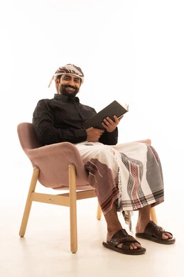 Looking at the camera with expressions of happiness and joy, the concept of self-learning, spending time reading, learning, and culture, a portrait of a Yemeni Arab man wearing a shirt and a sarong with a turban sitting on a chair reading a book, white background.