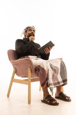 مفهوم التثقيف والبحث العلمي، قضاء وقت الفراغ بالتعلم والثقافة، هواية المطالعة والاستكشاف، بورتريه لرجل عربي يمني يرتدي القميص والازار مع العمامة يجلس على كرسي يمسك كتاب ويضع يده على ذقنه ينظر إلى شيء ما بتعابير التفكير، خلفية بيضاء