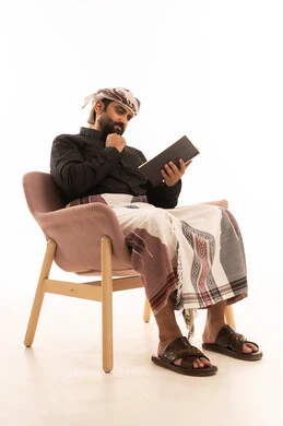 قضاء وقت الفراغ بالتعلم والثقافة، مفهوم التثقيف والبحث العلمي، هواية المطالعة والاستكشاف، بورتريه لرجل عربي يمني يرتدي القميص والازار مع العمامة يجلس على كرسي يمسك كتاب ويضع يده على ذقنه بتعابير الاندماج و التركيز، خلفية بيضاء