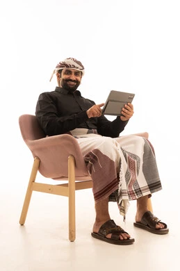 الدردشة مع الأهل والأصدقاء، استخدام التكنولوجيا و التقنيات الحديثة , بورتريه لرجل عربي يمني يرتدي القميص والازار مع العمامة يجلس على كرسي يحمل بيده جهاز التابليت، تصفح برامج التواصل الاجتماعي ، تطبيقات وبرامج الأجهزة اللوحية، خلفية بيضاء