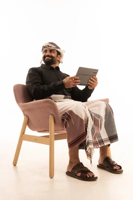 Looking at something, chatting with family and friends, using technology and modern techniques, a portrait of an Arab Yemeni man wearing a shirt and a waist wrap with a turban sitting on a chair holding a tablet, browsing social media programs, applications, and tablet programs, white background.