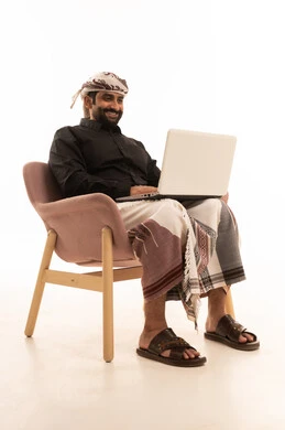 The development of modern devices and technologies, browsing the internet and social media programs, using a laptop to accomplish tasks, a portrait of an Arab Yemeni man wearing a shirt and a waist wrap with a turban sitting on a chair holding a laptop in his hands, white background.