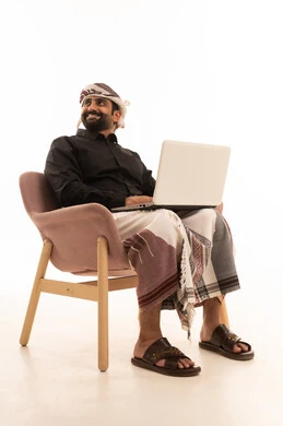استخدام الحاسب المحمول في انجاز المهام, تصفح الانترنت وبرامج التواصل الاجتماعي, بورتريه لرجل عربي يمني يرتدي القميص والازار مع العمامة يجلس على كرسي يمسك بيدية الابتوب, تطور الاجهزة والتقينات الحديثة, خلفية بيضاء