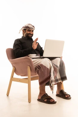 رفع كف اليد بتعابير السلام و التحية، اجراء مكالمة فيديو، العمل عن بعد، استخدام التقنية الحديثه المتطورة في الحياة العملية، بورتريه لرجل عربي يمني يرتدي القميص والازار مع العمامة يجلس على كرسي يقوم بالعمل على الحاسب المحمول، خلفية بيضاء