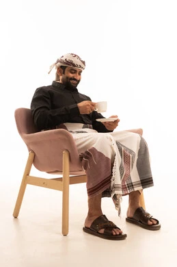 تأثير الكافيين على نشاط الدماغ والذاكرة، الاستمتاع بتناول المشروبات الساخنة والأعشاب الشتوية الدافئة، بورتريه لرجل عربي يمني يرتدي القميص والازار مع العمامة يجلس على كرسي يحمل بيده كوب من القهوة، خلفية بيضاء