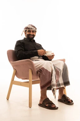الاستمتاع بتناول المشروبات الساخنة والأعشاب الشتوية الدافئة، تأثير الكافيين على نشاط الدماغ والذاكرة، بورتريه لرجل عربي يمني يرتدي القميص والازار مع العمامة يجلس على كرسي يحمل بيده كوب من القهوة، خلفية بيضاء
