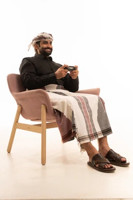 Spending enjoyable time playing video games, enjoying modern technologies and gadgets, the concept of fun and entertainment, gestures of immersion and interaction, a portrait of a Yemeni Arab man wearing a shirt and a sarong with a turban sitting on a chair holding a game controller, white background.