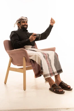 Spending enjoyable time at home, fun, entertainment, and interaction, home recreational activities, using modern technology for entertainment, a portrait of an Arab Yemeni man wearing a shirt and a sarong with a turban sitting on a chair holding a game controller raising his hand in gestures of victory, playing video games with gestures of joy and happiness, white background.