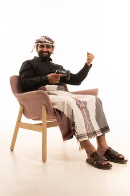 Fun, entertainment, and interaction, home recreational activities, spending enjoyable time at home, using modern technology for entertainment, a portrait of a Yemeni Arab man wearing a shirt and a sarong with a turban sitting on a chair holding a game controller raising his hand in victory gestures, playing video games with gestures of joy and happiness, white background.