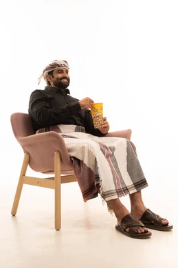 إيماءات السعادة والمتعة، الاستعداد لمشاهدة الافلام وإحضار السناكات اللذيذة، مشاهدة فلم سينمائي، بورتريه لرجل عربي يمني يرتدي القميص والازار مع العمامة يجلس على كرسي يحمل بيده علبة الفوشار، خلفية بيضاء