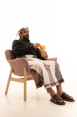 إيماءات الاندماج و الاندهاش، الاستعداد لمشاهدة الافلام وإحضار السناكات اللذيذة، مشاهدة فلم سينمائي، بورتريه لرجل عربي يمني يرتدي القميص والازار مع العمامة يجلس على كرسي يحمل بيده علبة الفوشار، خلفية بيضاء