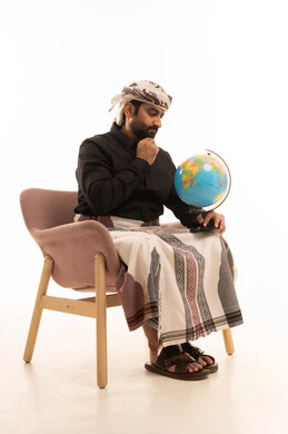 Distinguishing the land and maritime borders of countries, looking at something, recognizing the geography of the country, a love for research and exploration, a portrait of an Arab Yemeni man wearing a shirt and a sarong with a turban sitting on a chair holding a small model of the globe in his hand and resting his hands on his chin, white background.