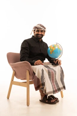 Recognizing the geography of the country, a love for research and exploration, distinguishing the land and maritime borders of countries, a portrait of a Yemeni Arab man wearing a shirt and a sarong with a turban sitting on a chair holding a miniature globe in his hand with gestures of happiness and joy, white background.