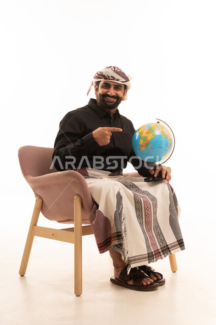 The love of research and exploration, getting to know the geography of countries, distinguishing the land and maritime borders of nations, a portrait of an Arab Yemeni man wearing a shirt and a sarong with a turban sitting on a chair holding a miniature globe in his hand and pointing to it with gestures of happiness and joy, white background.