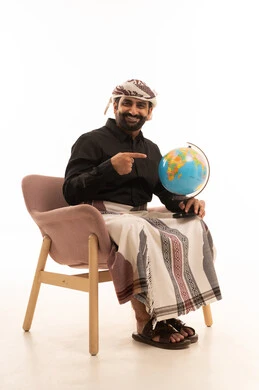 The love of research and exploration, getting to know the geography of countries, distinguishing the land and maritime borders of nations, a portrait of an Arab Yemeni man wearing a shirt and a sarong with a turban sitting on a chair holding a miniature globe in his hand and pointing to it with gestures of happiness and joy, white background.