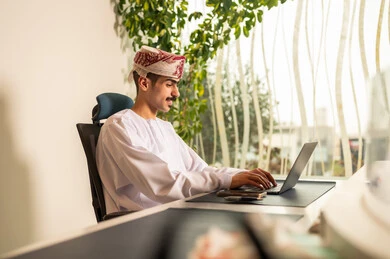 مفهوم التركيز و الدقة في الإنجاز، مهنة و وظيفة عمانية مكتبية, اجتماع عمل عن بعد ، استخدام الحاسب و التقنية في مجال الاعمال، إتمام مهام العمل في المكتب، موظف عربي خليجي عماني يرتدي الدشداشة والكمة ويقوم باستخدام اللابتوب