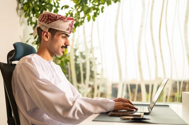 موظف عربي خليجي عماني يرتدي الدشداشة والكمة ويقوم باستخدام اللابتوب، اجتماع عمل عن بعد ، استخدام الحاسب و التقنية في مجال الاعمال، مفهوم التركيز و الدقة في الإنجاز، مهنة و وظيفة عمانية مكتبية, إتمام مهام العمل في المكتب
