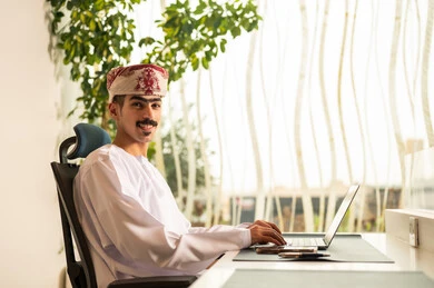 Omani office profession and job, remote work meeting, using computers and technology in the field of business, the concept of focus and accuracy in achievement, completing office tasks, an Arab Gulf Omani employee wearing a dishdasha and kumma using a laptop.
