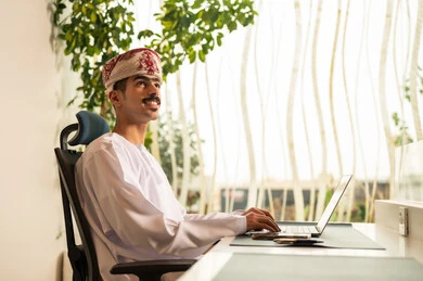 موظف عربي خليجي عماني يرتدي الدشداشة والكمة ويقوم باستخدام اللابتوب، اجتماع عمل عن بعد ، استخدام الحاسب و التقنية في مجال الاعمال، مفهوم التركيز و الدقة في الإنجاز، مهنة و وظيفة عمانية مكتبية, إتمام مهام العمل في المكتب