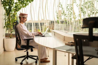 اجتماع عمل عن بعد ، استخدام الحاسب و التقنية في مجال الاعمال، مفهوم التركيز و الدقة في الإنجاز، مهنة و وظيفة عمانية مكتبية, إتمام مهام العمل في المكتب، موظف عربي خليجي عماني يرتدي الدشداشة والكمة ويقوم باستخدام اللابتوب