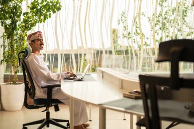 مهنة و وظيفة عمانية مكتبية, اجتماع عمل عن بعد ، استخدام الحاسب و التقنية في مجال الاعمال، مفهوم التركيز و الدقة في الإنجاز، إتمام مهام العمل في المكتب، موظف عربي خليجي عماني يرتدي الدشداشة والكمة ويقوم باستخدام اللابتوب