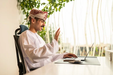 Remote work meeting, using computers and technology in the field of business, the concept of focus and precision in achievement, an Omani office profession and job, completing tasks in the office, an Arab Gulf Omani employee wearing a dishdasha and kumma using a laptop.