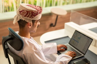 استخدام الحاسب و التقنية في مجال الاعمال، اجتماع عمل عن بعد ، مفهوم التركيز و الدقة في الإنجاز، مهنة و وظيفة عمانية مكتبية, إتمام مهام العمل في المكتب، موظف عربي خليجي عماني يرتدي الدشداشة والكمة ويقوم باستخدام اللابتوب