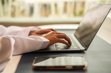 Remote work meeting, using computers and technology in the field of business, the concept of focus and precision in achievement, an Omani office profession and job, completing tasks in the office, an Arab Gulf Omani employee wearing a dishdasha and kumma using a laptop.